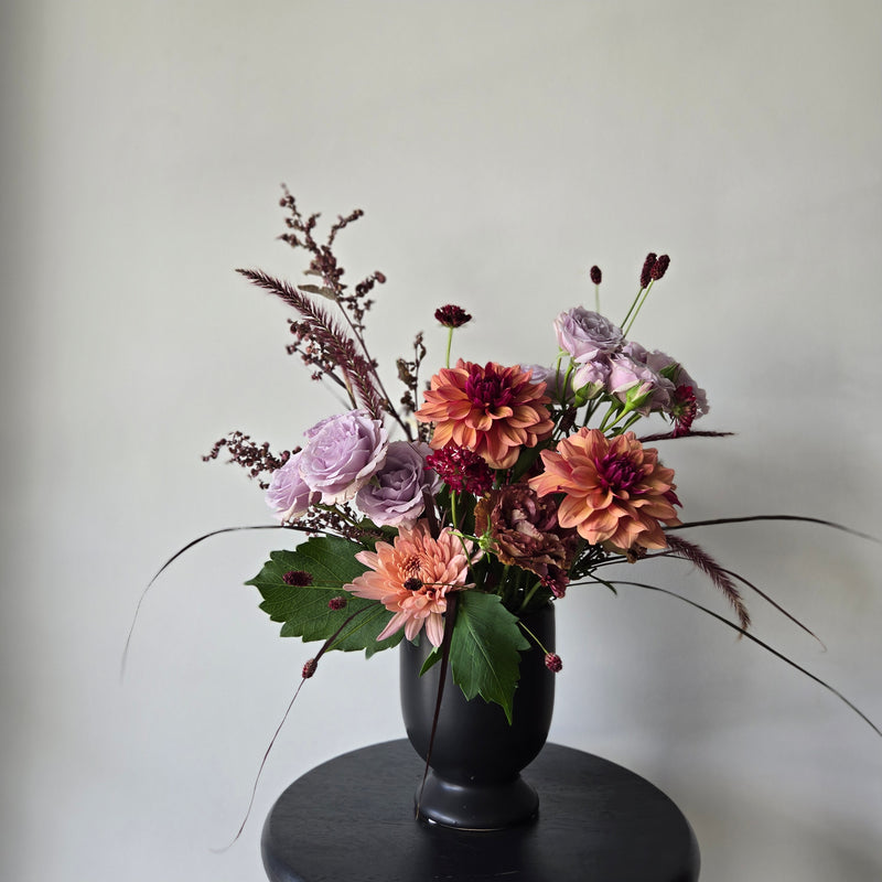 Floral arrangement with pink, purple, and green flowers on a neutral background in a black pedestal vase